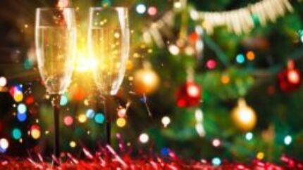 Two champagne glasses with festive Christmas decorations and lights in the background.